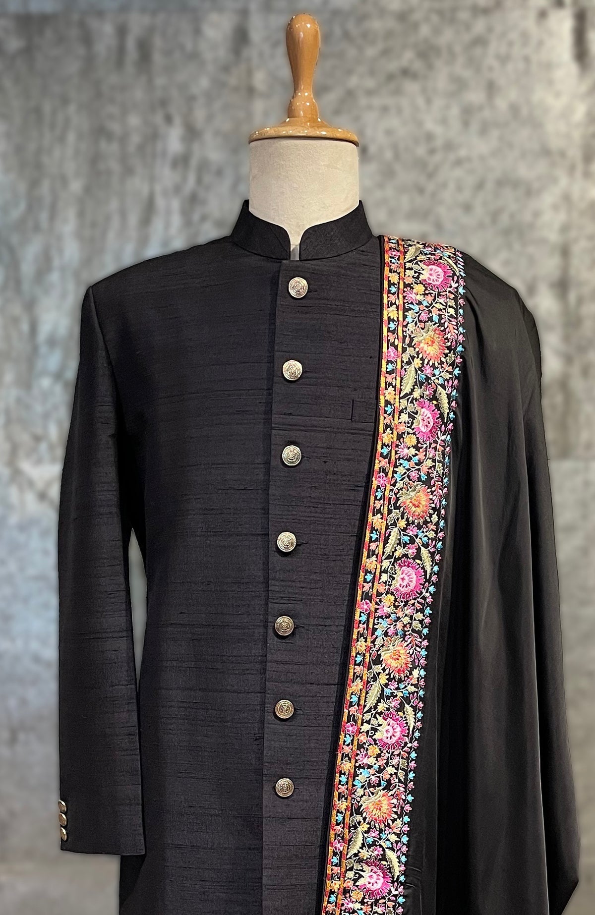 Black sherwani with gold buttons and a colorful floral embroidered shawl.