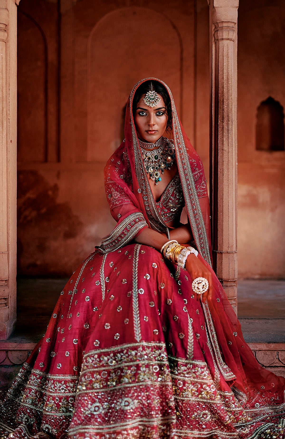 A woman in a red embroidered lehenga set with heavy jewelry poses in a traditional setting.