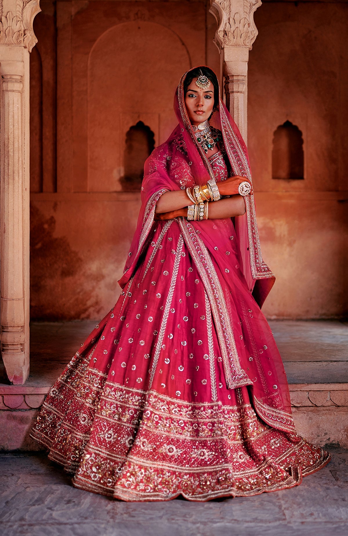 A model poses in a red raw silk lehenga set with intricate embroidery and a sheer organza dupatta.