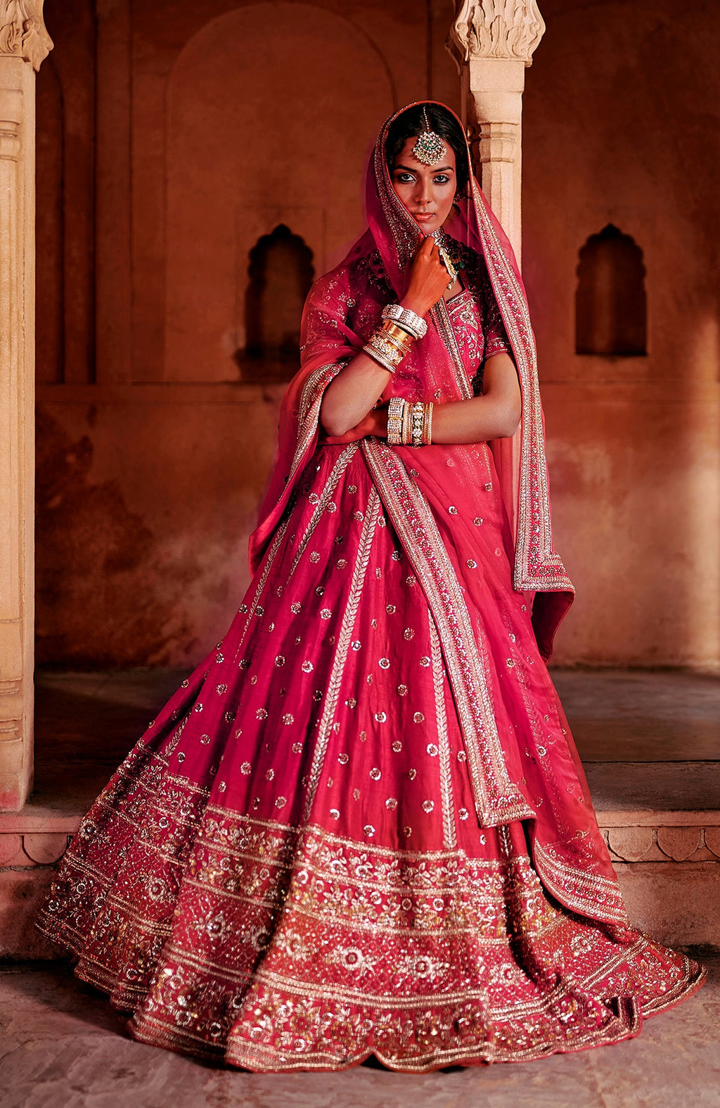A model in a red bridal lehenga set with intricate gold embroidery and heavy jewelry.