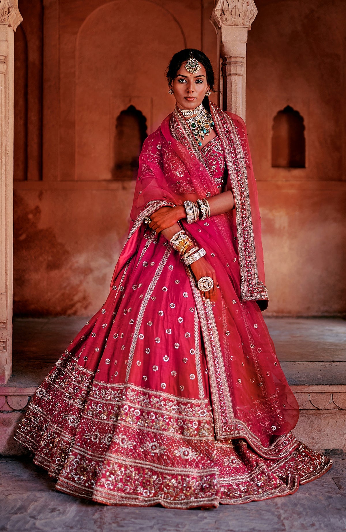 A woman models a red bridal lehenga set with intricate embroidery and jewelry.