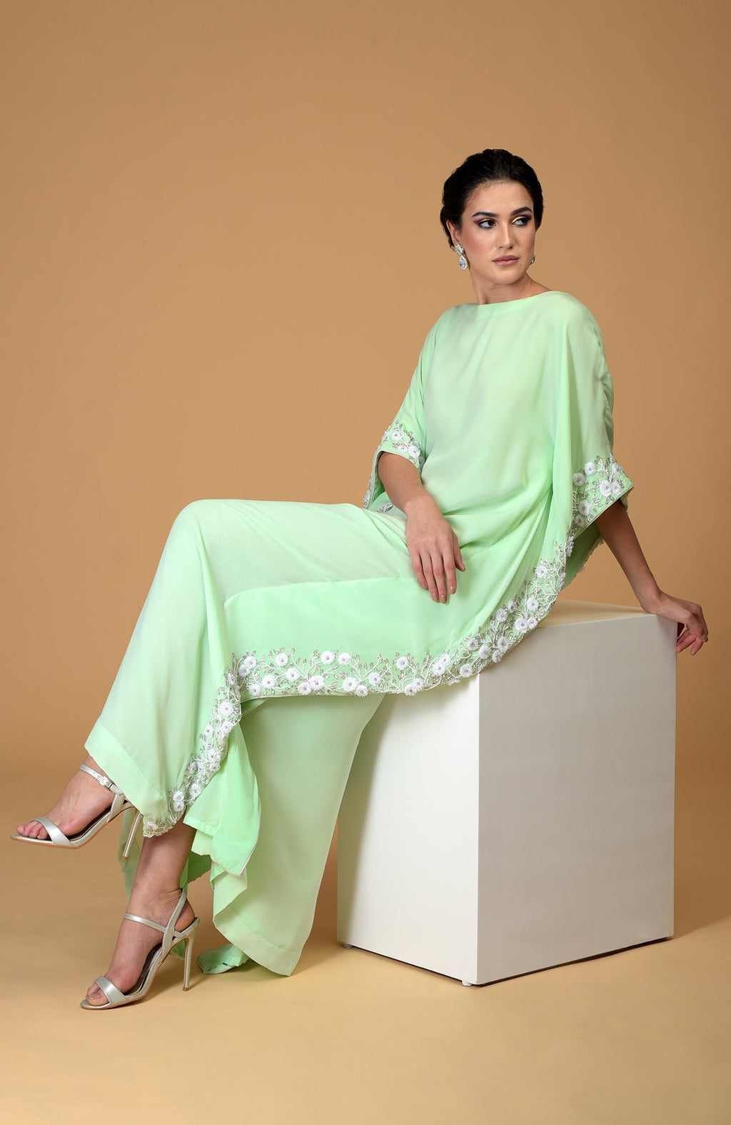 A woman in a mint green floral hand-embroidered dress sits on a white cube.