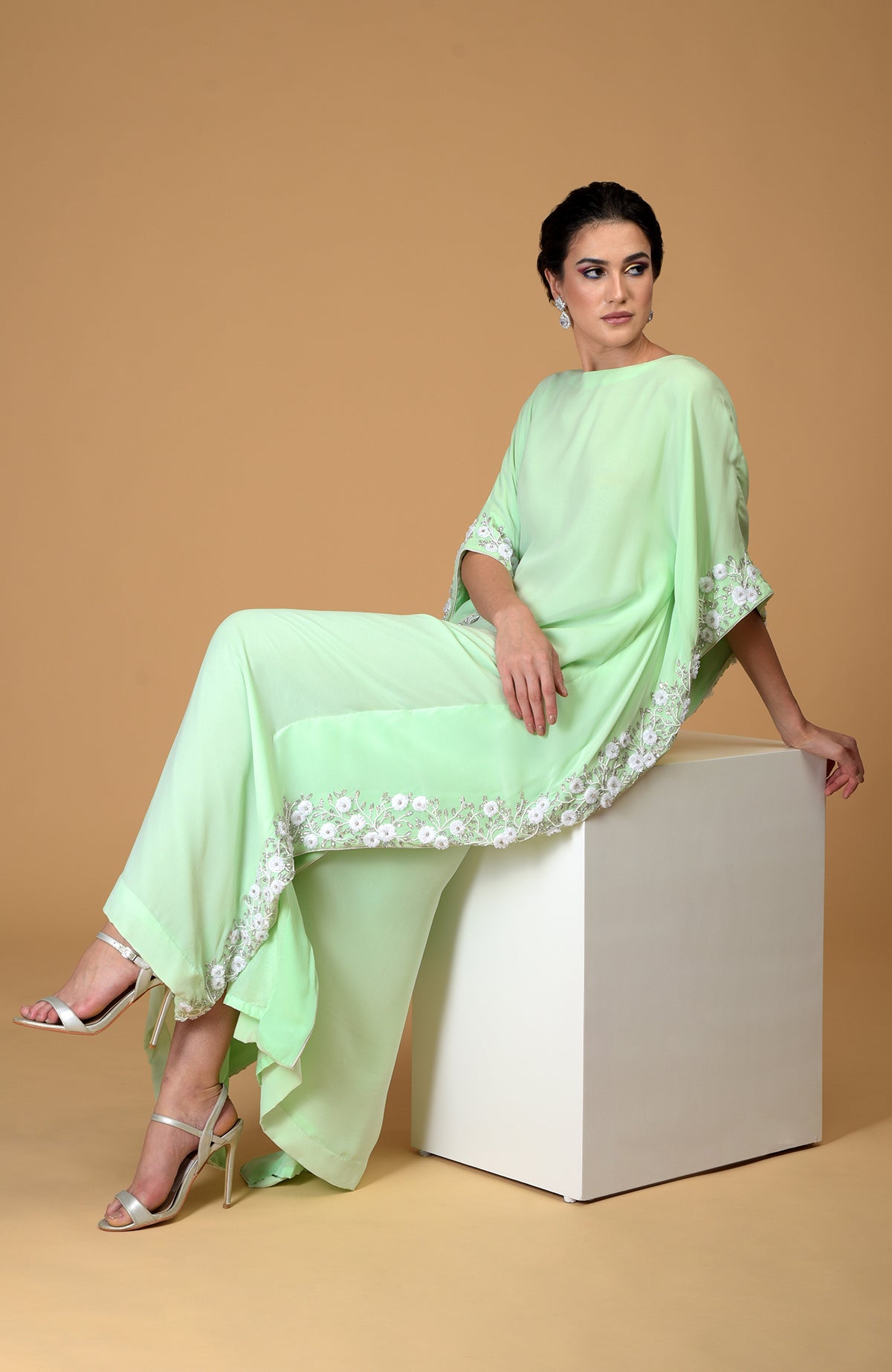 A woman in a mint green floral hand-embroidered dress sits on a white cube.