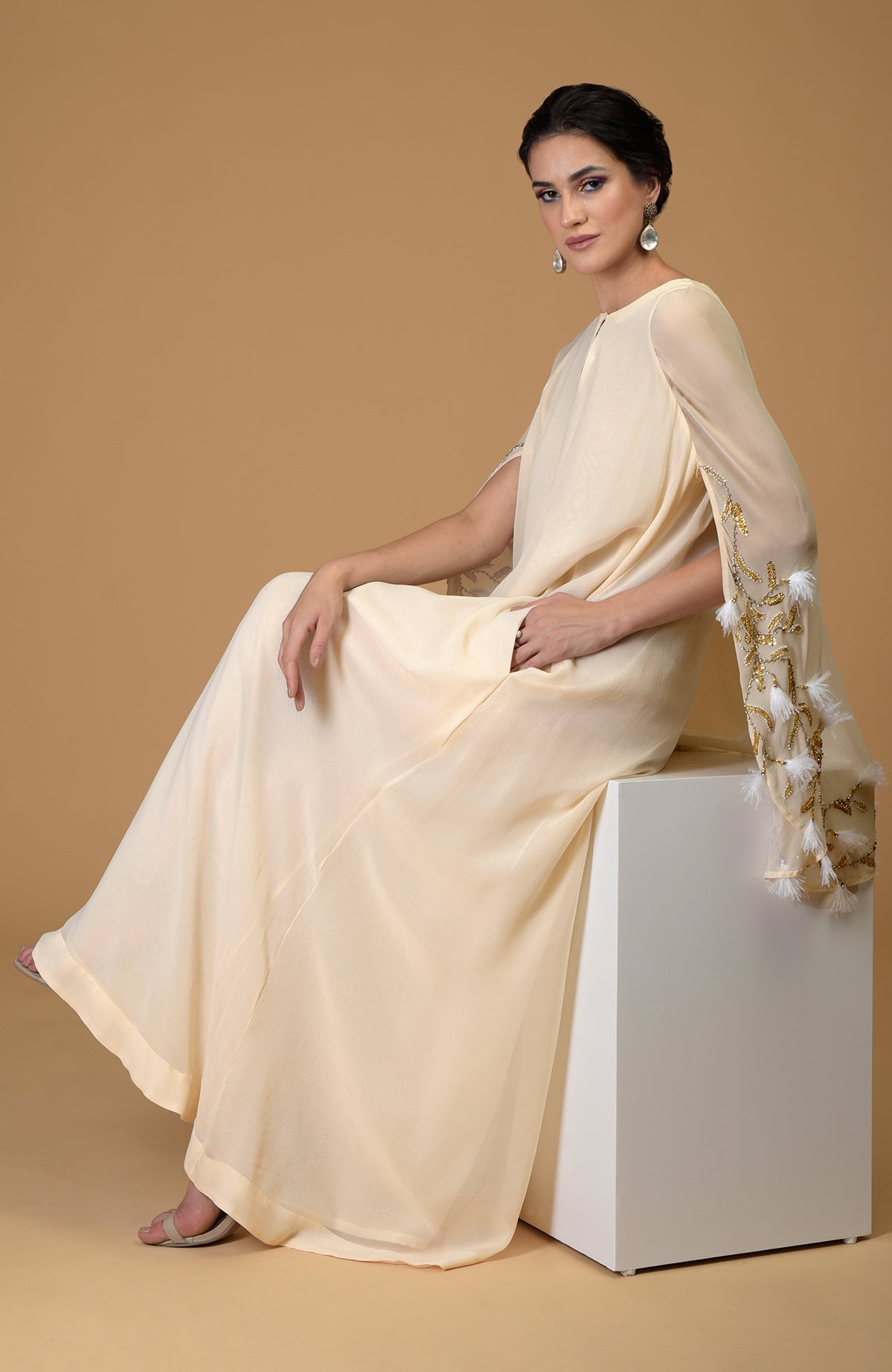 A woman in a cream cape dress with gold and white feather embroidery on the sleeves.