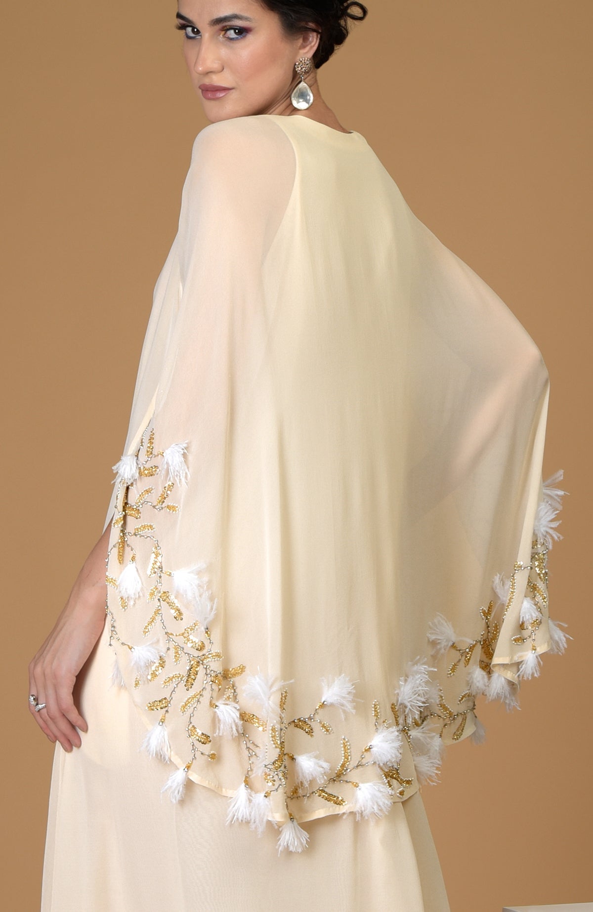 A woman models a cream-colored cape dress with gold and white feather embroidery.