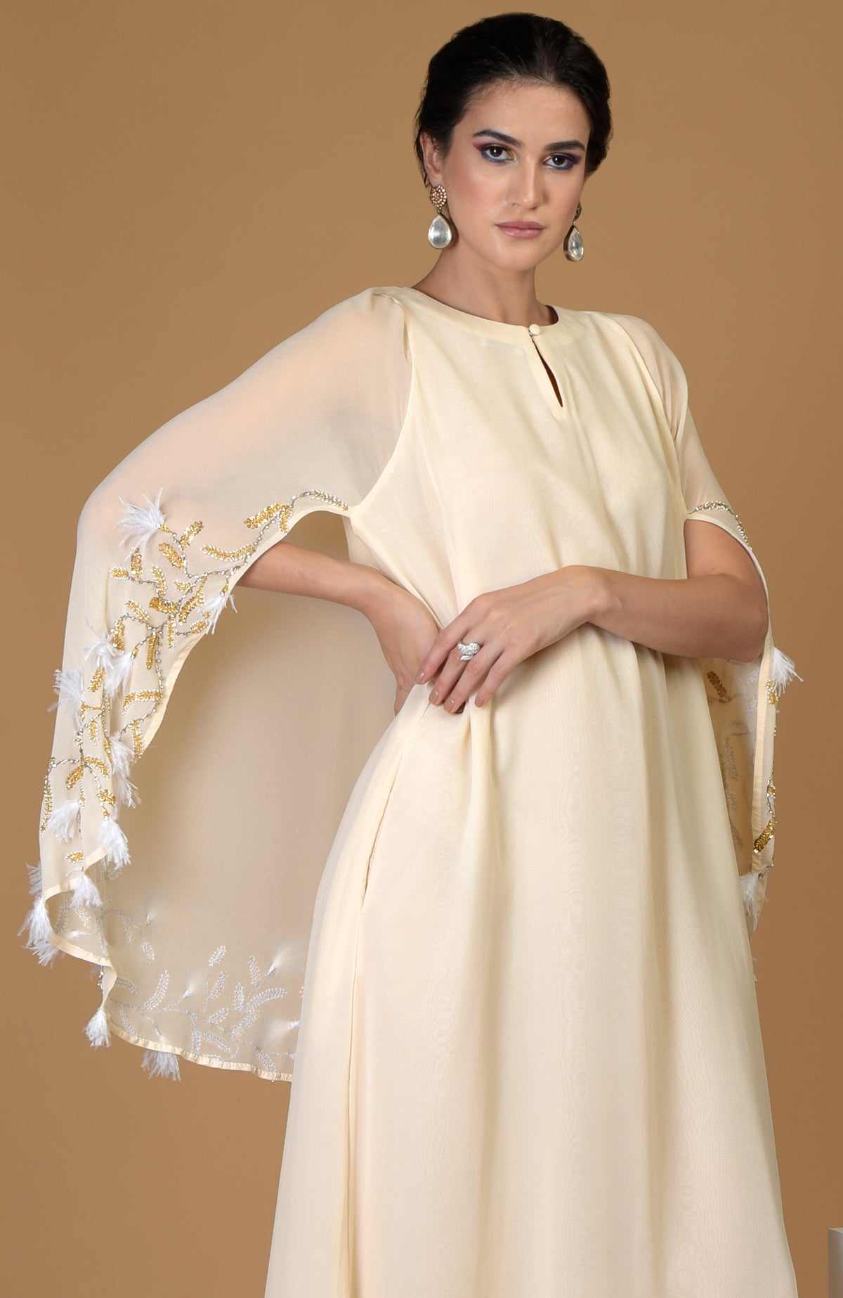 A woman models a cream-colored dress with a cape featuring gold and white feather embroidery.