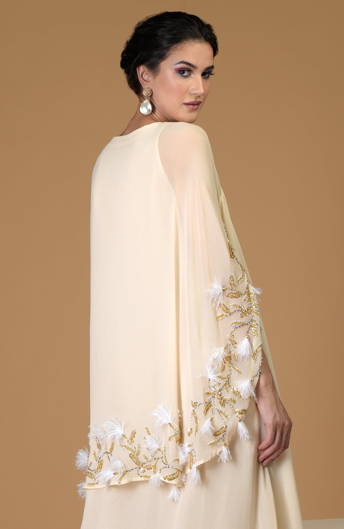 A woman models a cream-colored cape dress with gold and white feather embroidery.