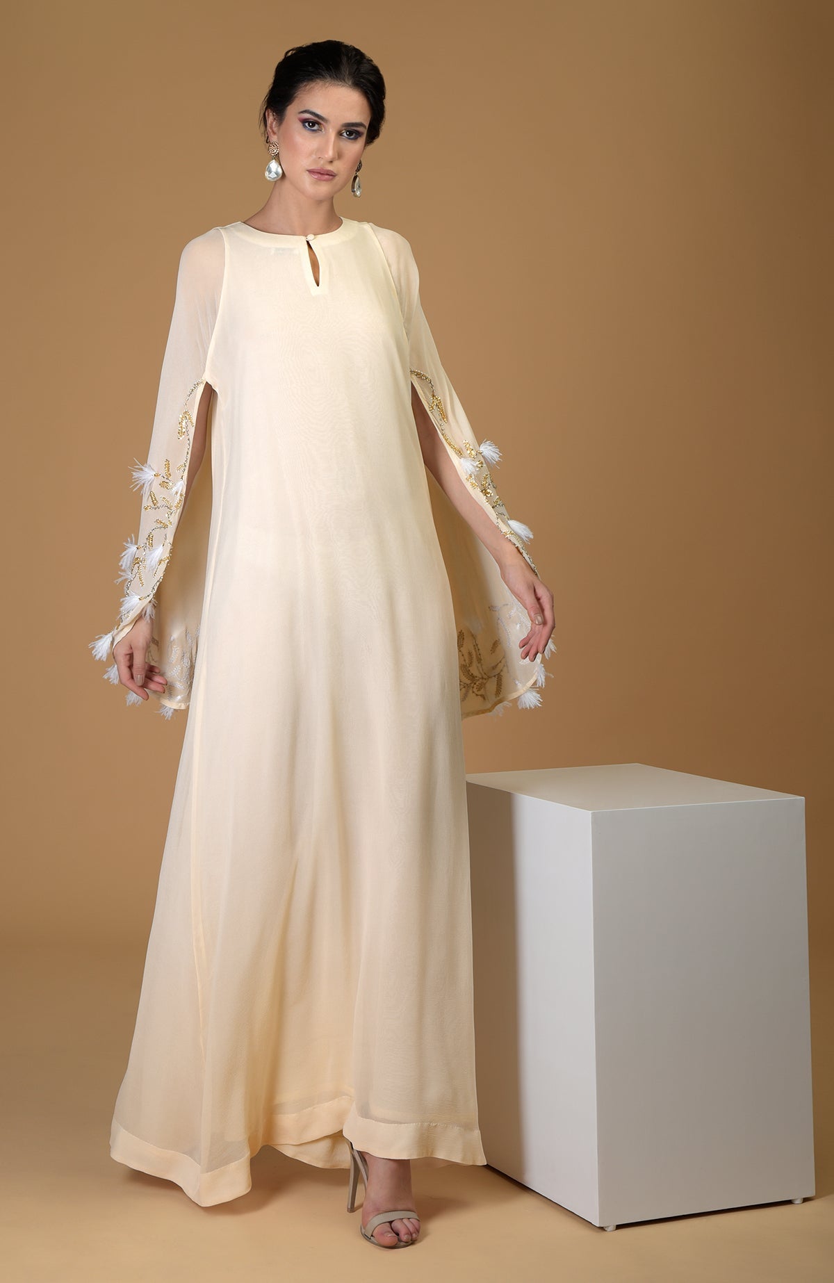 A model wears a cream-colored, hand-embroidered cape dress with feather and gold detailing.