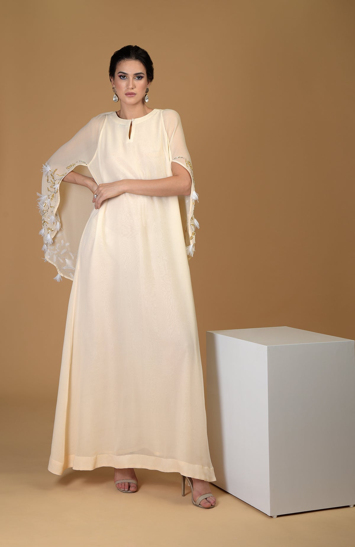A woman models a cream-colored, hand-embroidered cape dress with feather details.