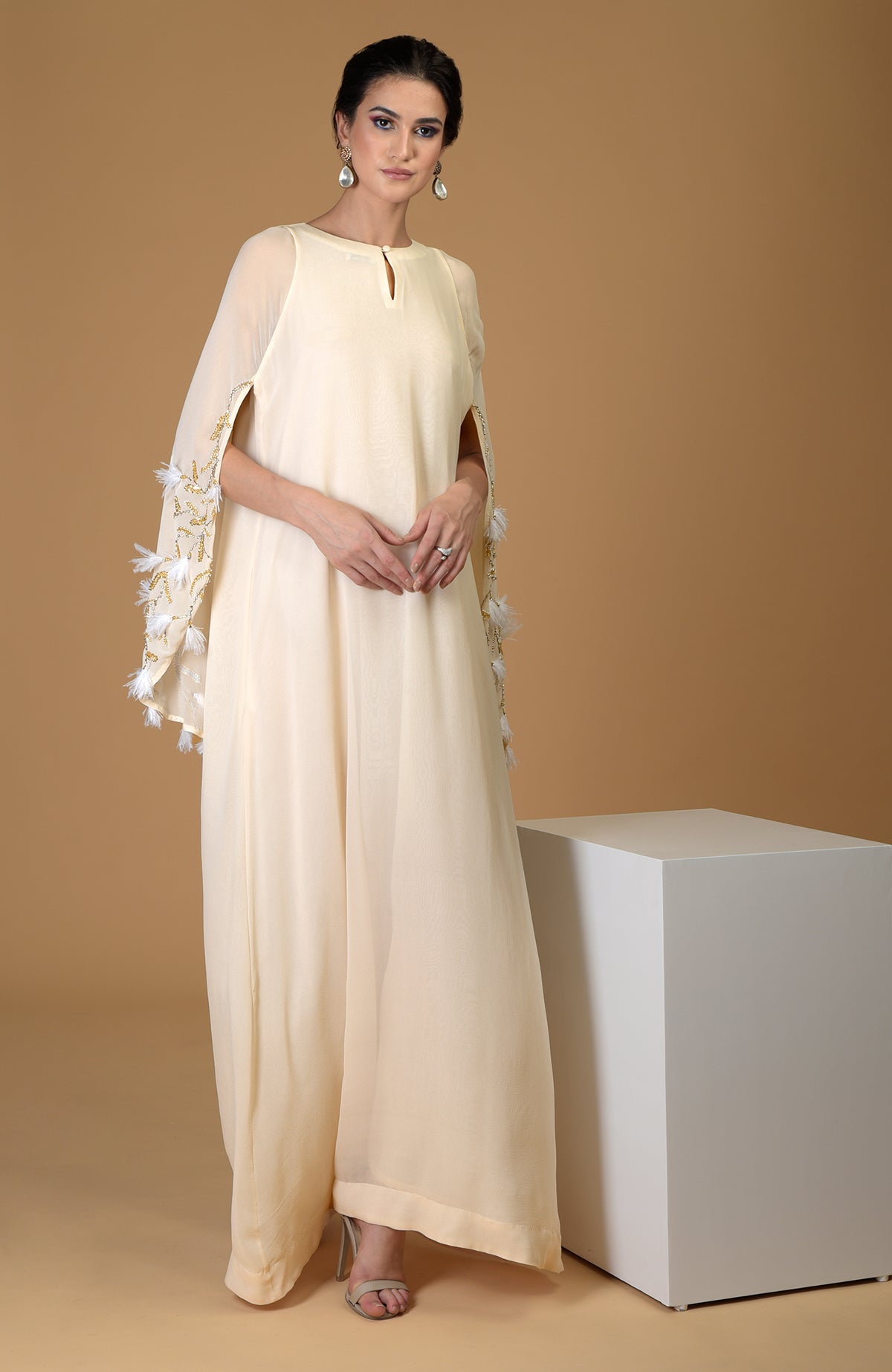 A woman models a cream-colored cape dress with gold embroidery and white feather details.