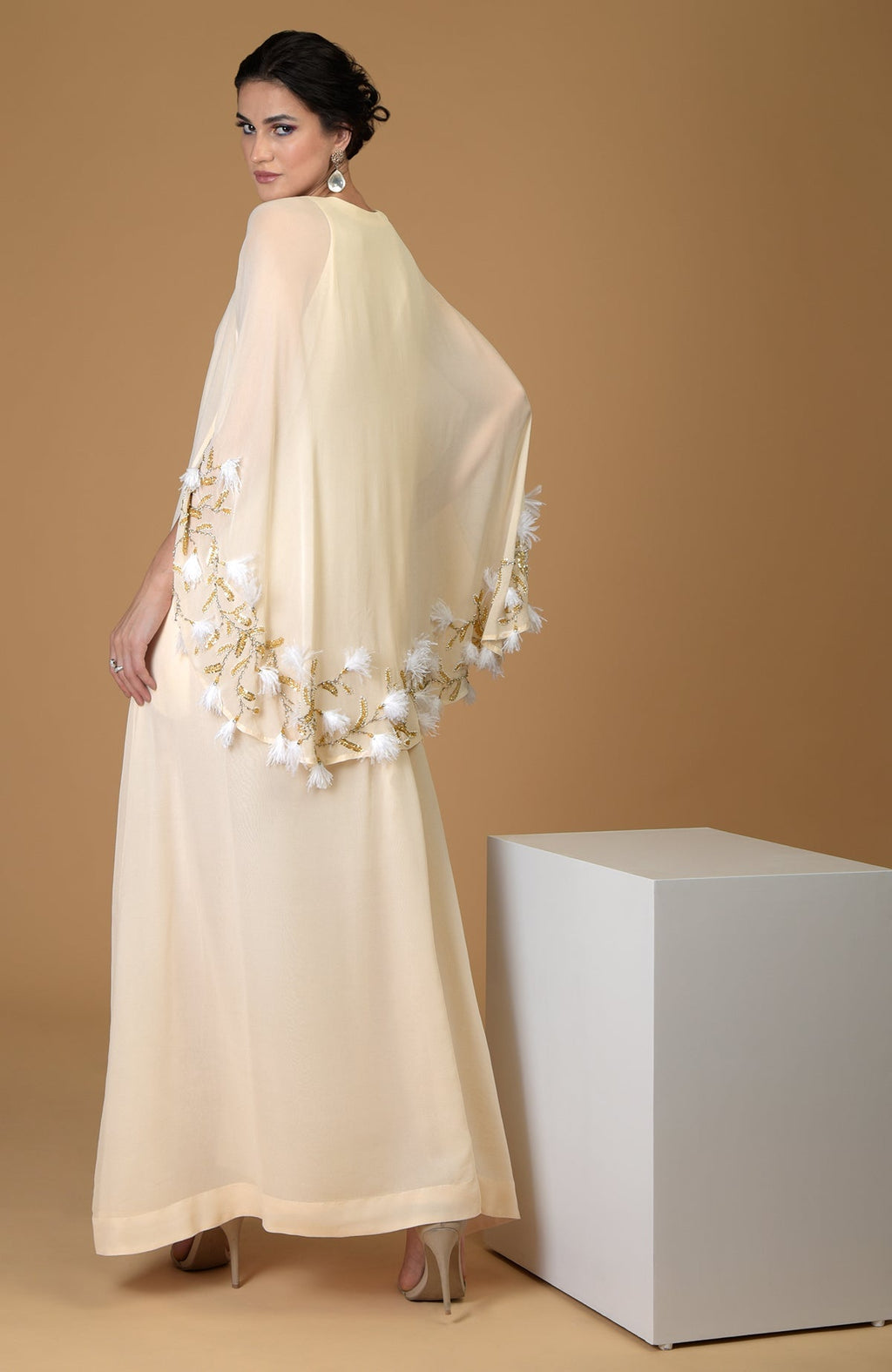 A woman models a cream-colored cape dress with gold and white feather embroidery.