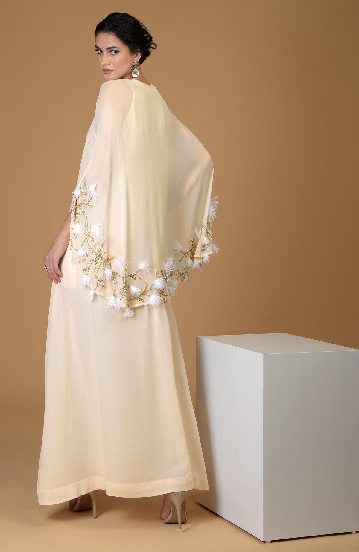 A woman models a cream-colored cape dress with gold and white feather embroidery.