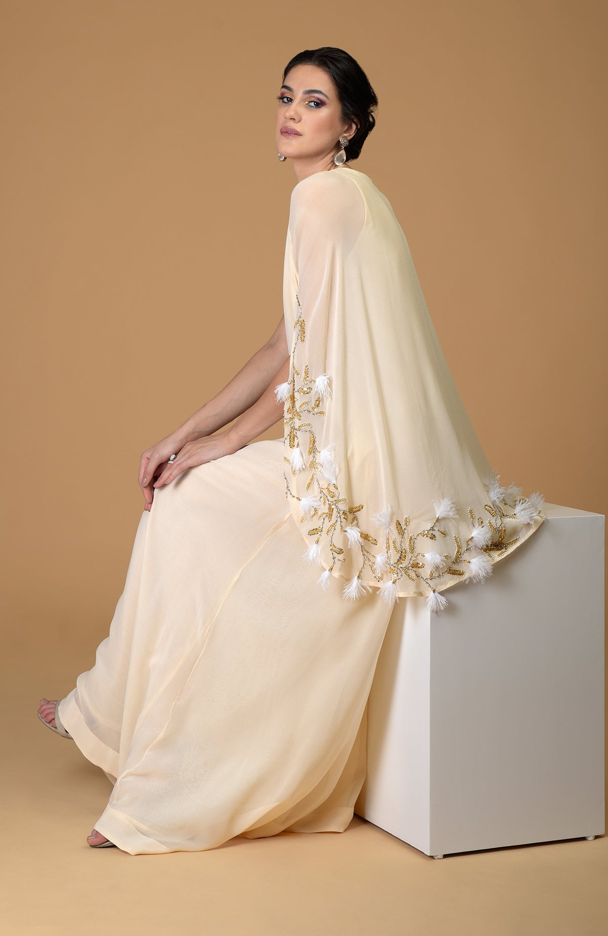 A woman in a cream cape dress with gold and white feather embroidery.