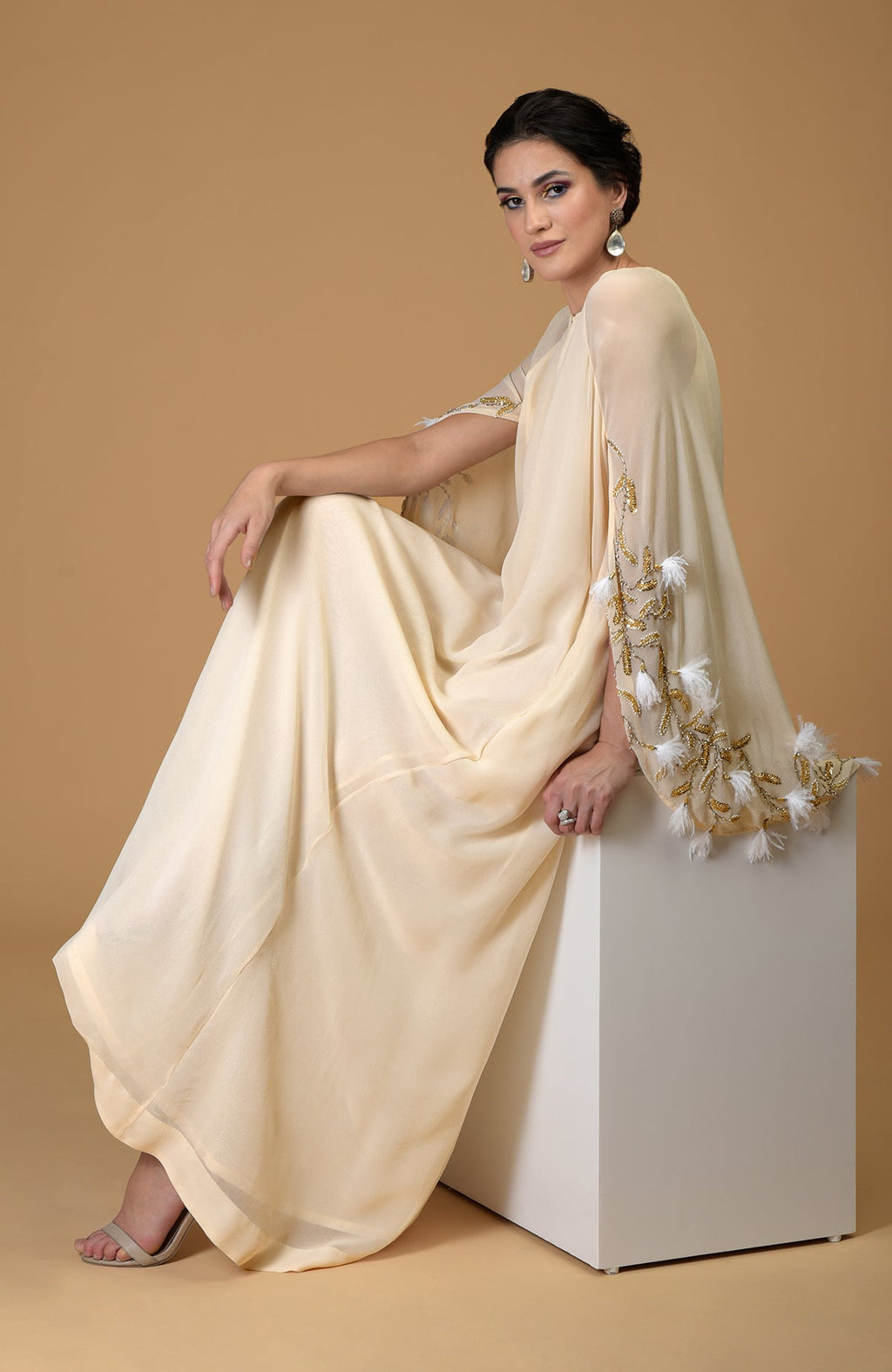 A woman models a hand-embroidered cream cape dress with feather and gold accents.