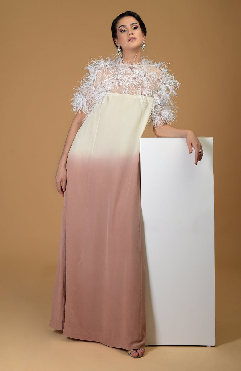 A woman in an ombre dress with a feather-embroidered sheer top leans on a white block.