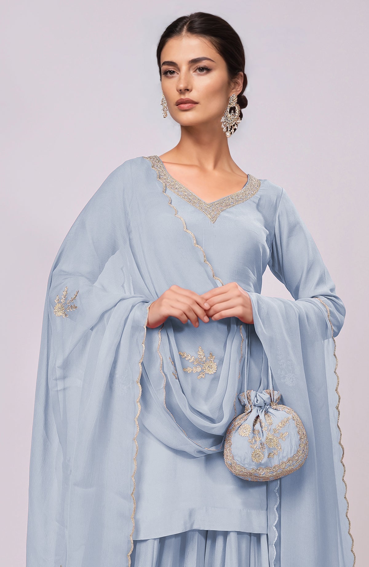 A woman in a dusty blue sharara set with gold embroidery and a matching potli bag.