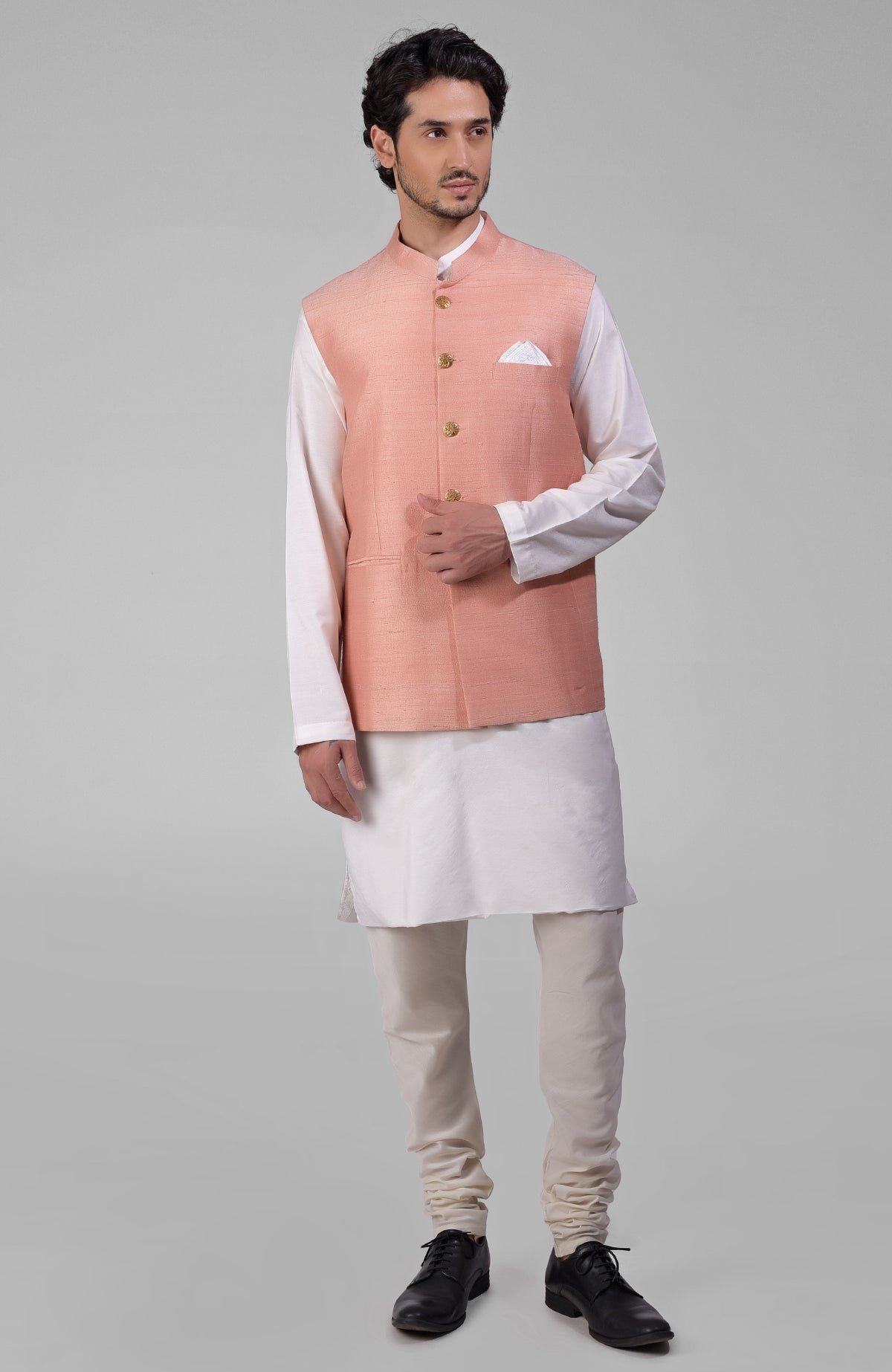 Peach raw silk waistcoat with gold buttons over a white kurta and light-colored pants.