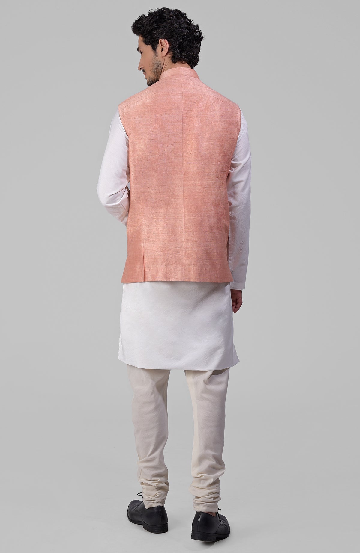 A man from behind wears a peach raw silk waistcoat over a white kurta and churidar.