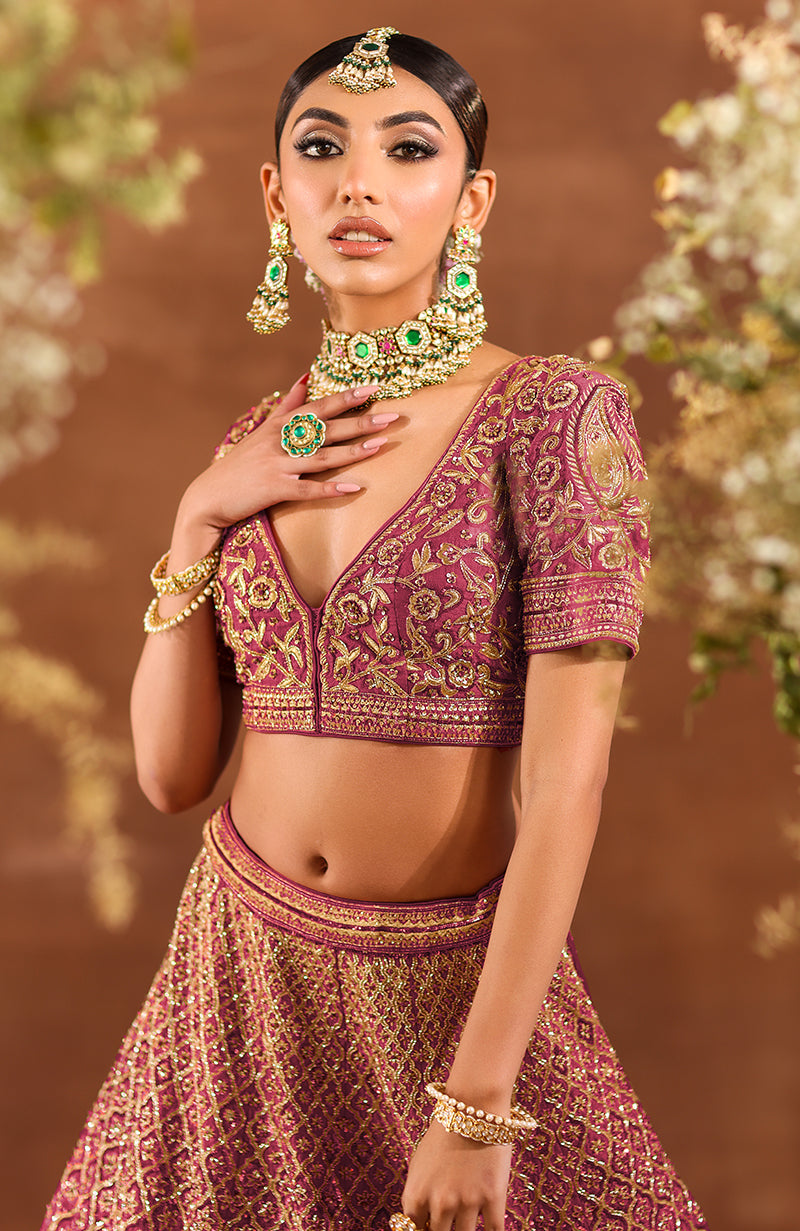 A model wears a deep rosewood lehenga set with intricate gold embroidery and green jewelry.