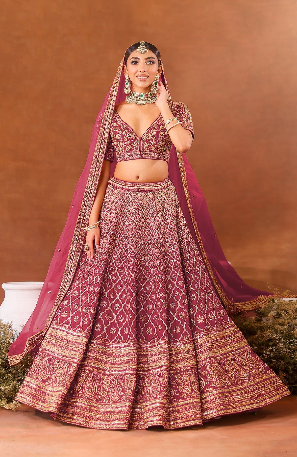 A woman models a purple wine lehenga set with gold embroidery and green jewelry.