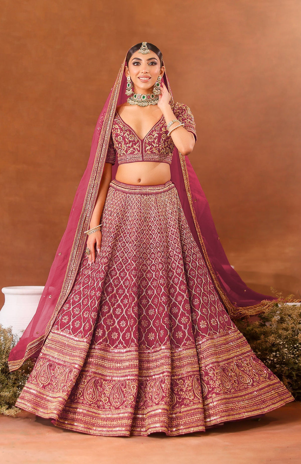 A woman models a deep rosewood lehenga set with gold embroidery and green jewelry.