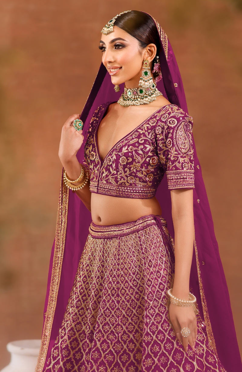 A woman in a purple wine lehenga set with gold embroidery and green jewelry.