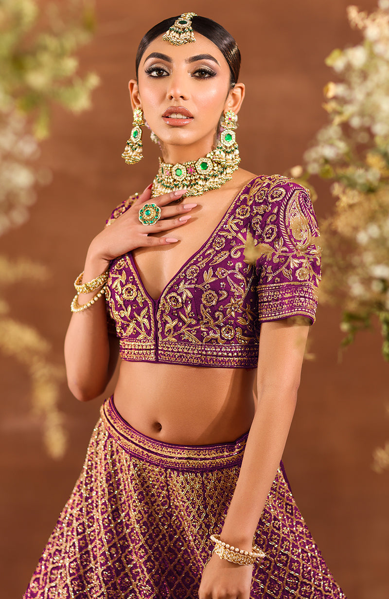 A woman models a purple wine lehenga set with gold embroidery and traditional jewelry.
