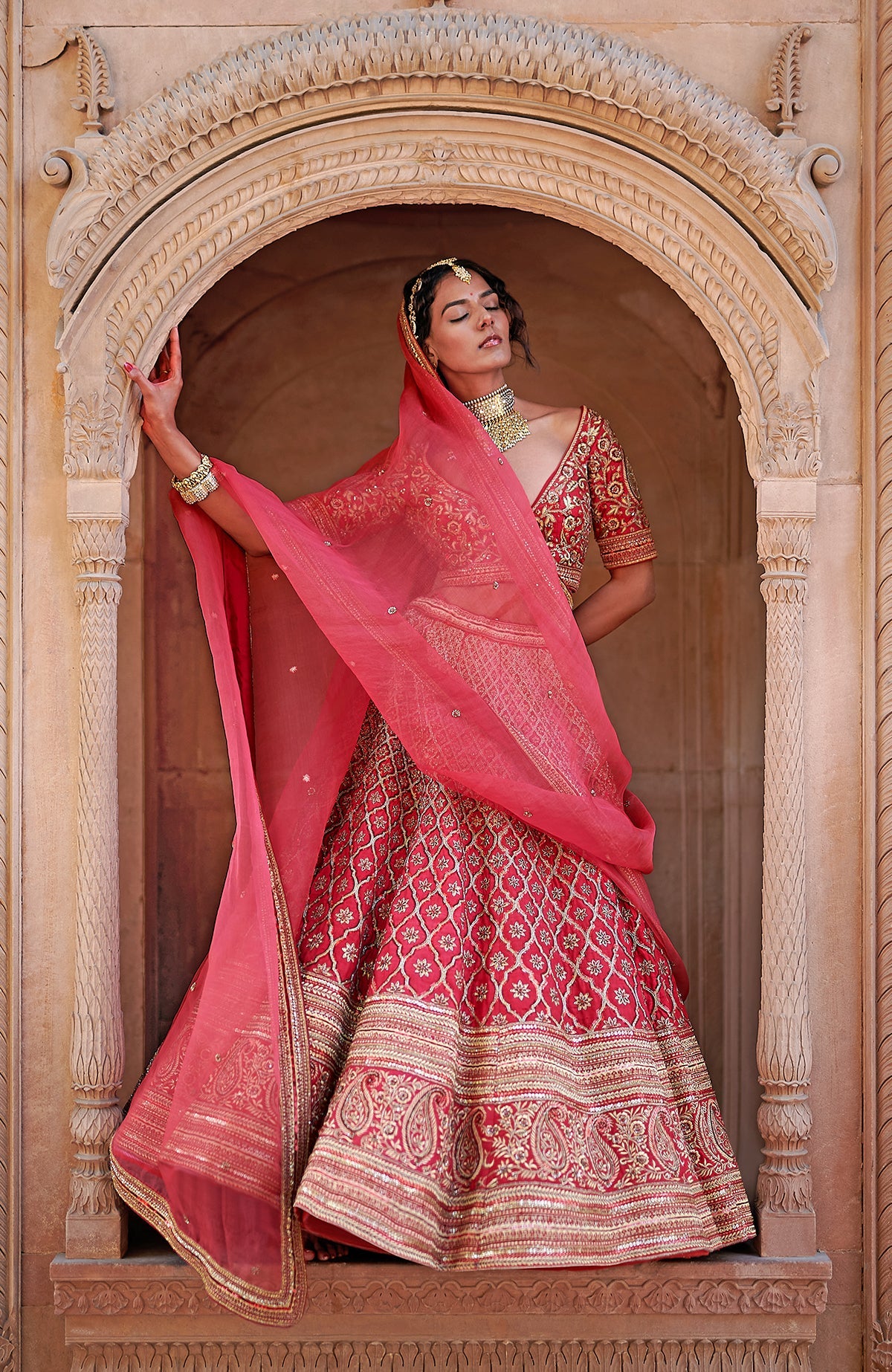 A model poses in a scarlet red bridal lehenga set with intricate gold embroidery.