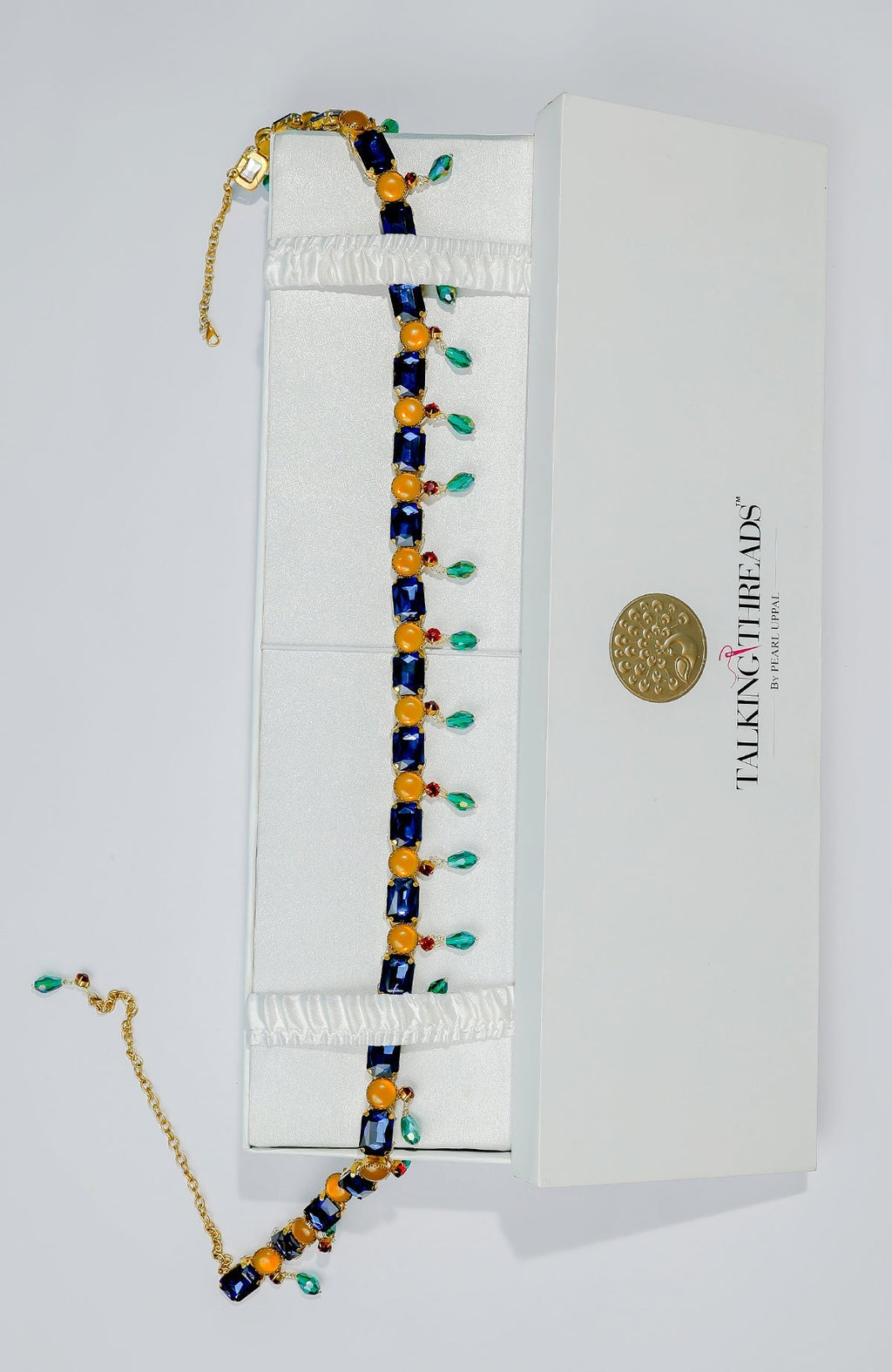 Blue jeweled crystal belt with orange accents and green dangling stones in a white box.