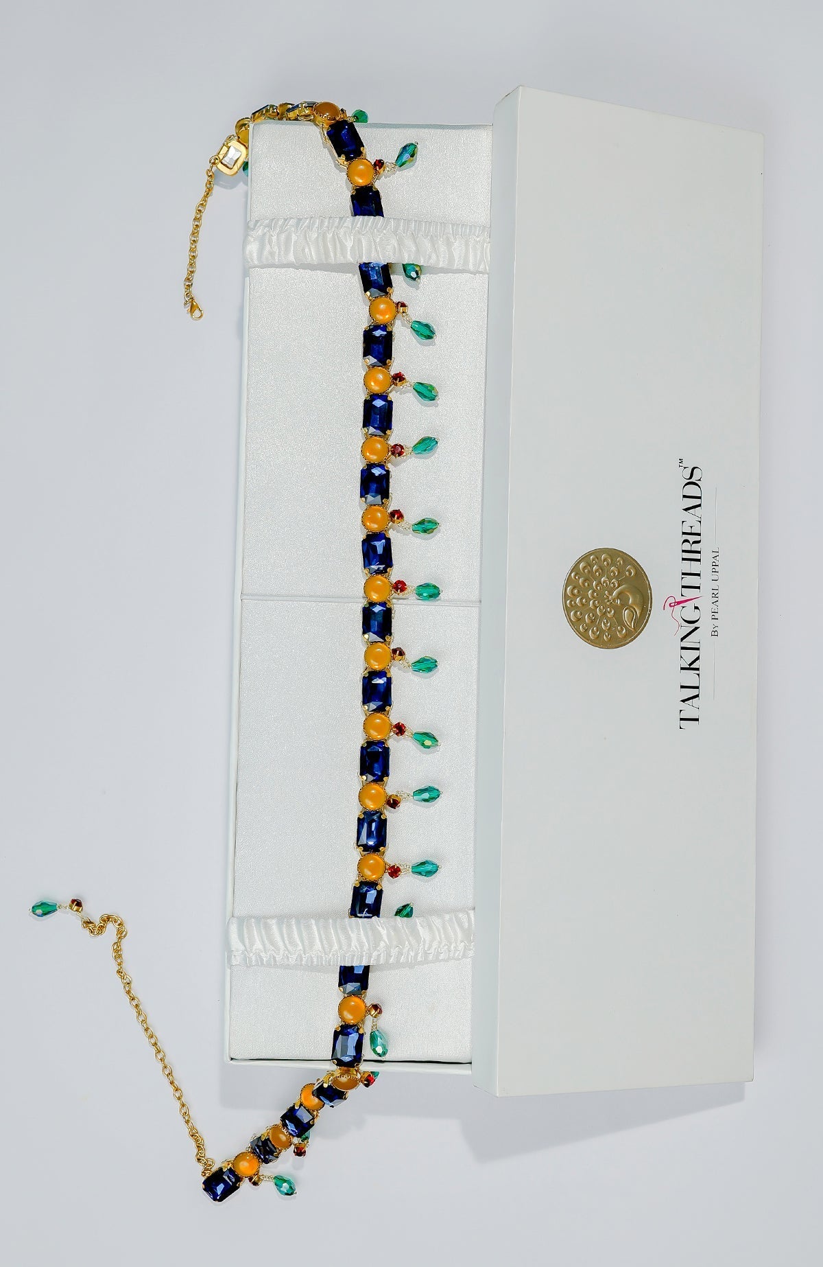 Blue jeweled crystal belt with orange accents and green dangling stones in a white box.