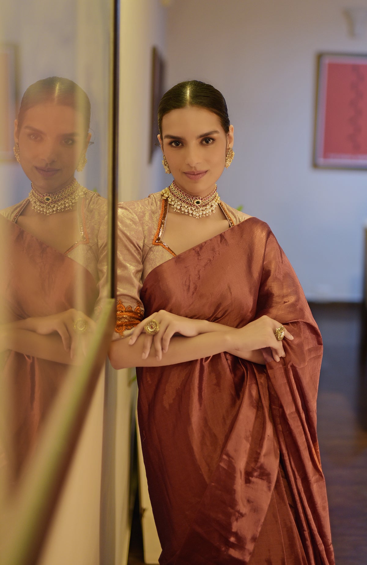 A woman in a copper tissue saree and zardozi embroidered blouse stands next to a reflection.