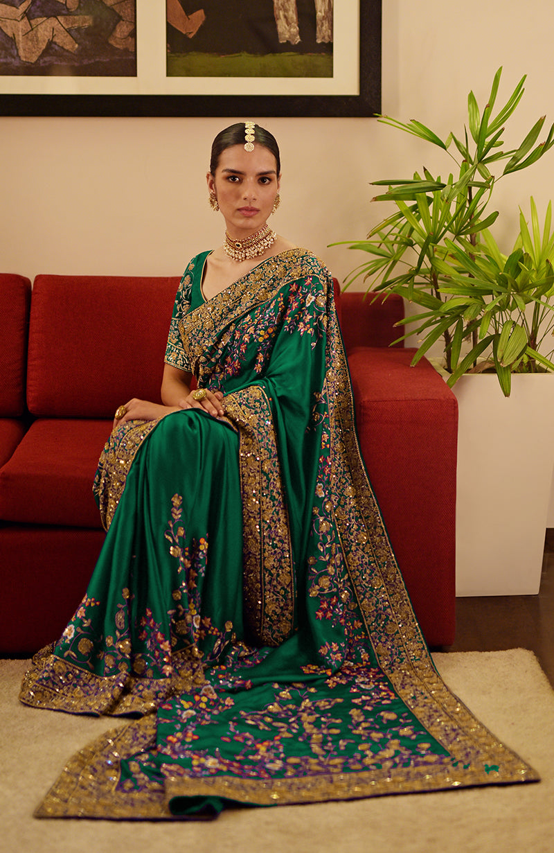 A woman sits on a red couch wearing a green embroidered saree with gold borders.