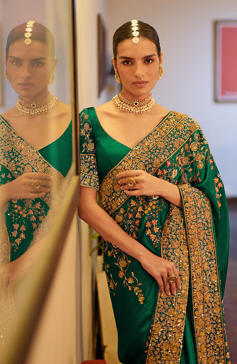 A woman in a deep emerald green saree with intricate gold and multi-color embroidery.