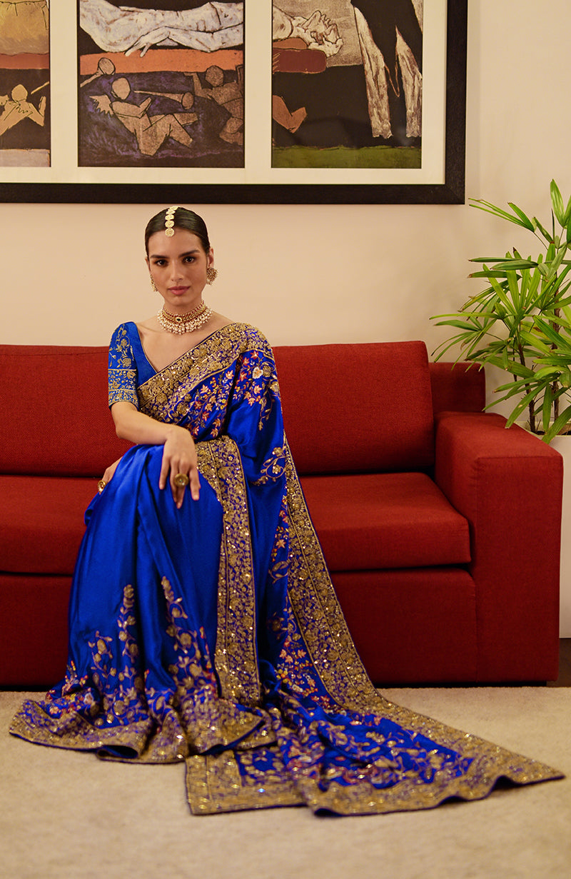 A woman in a royal blue saree with gold and multicolored embroidery sits on a red couch.
