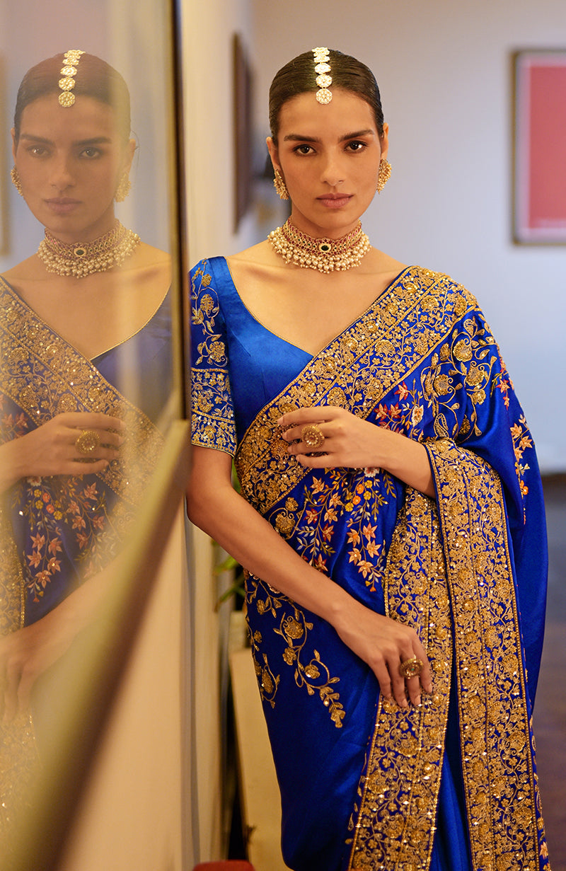A woman in a royal blue saree with gold and multicolored embroidery stands next to a mirror.
