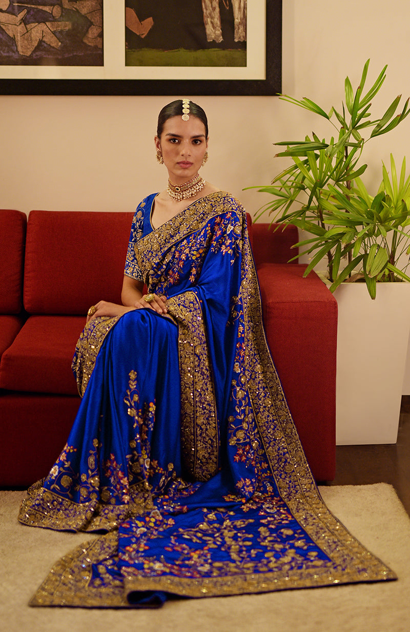 A woman sits on a red couch wearing a blue saree with gold and multicolor embroidery.