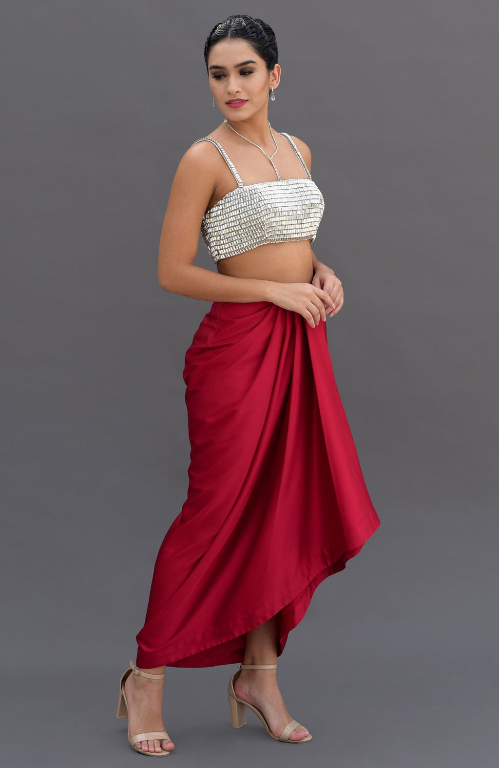 A woman models a silver crystal bustier and a red draped skirt with nude heels.