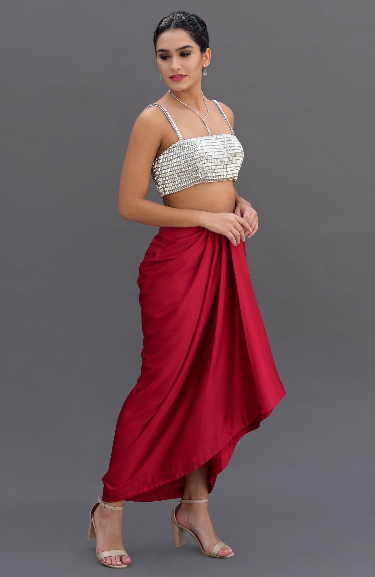 A woman models a silver crystal bustier and a red draped skirt with nude heels.