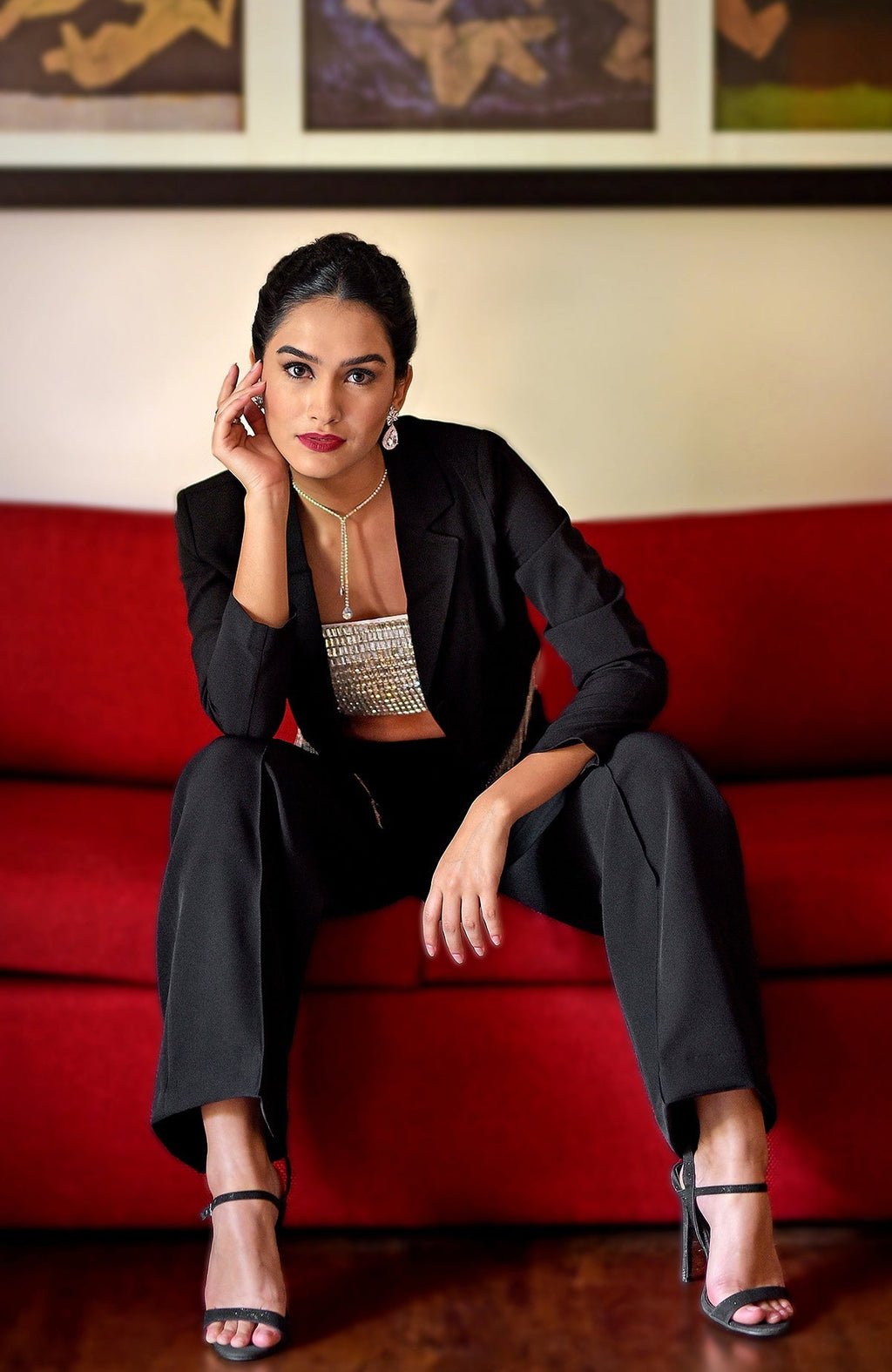 A woman in a black blazer, crystal bustier, and black pants sits on a red couch.