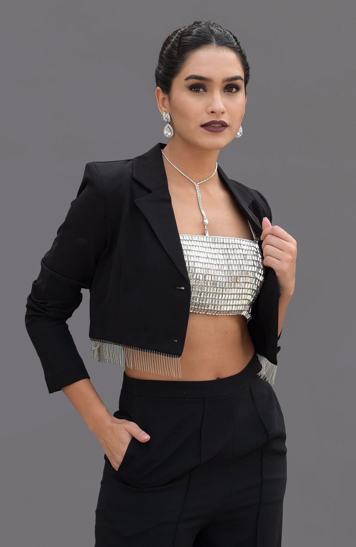 A woman models a black cropped blazer with fringe, a silver bustier, and black pants.