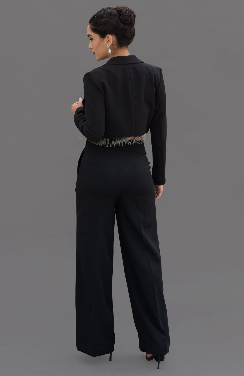 A woman in a black blazer with crystal fringe and matching pants stands with her back to the camera.