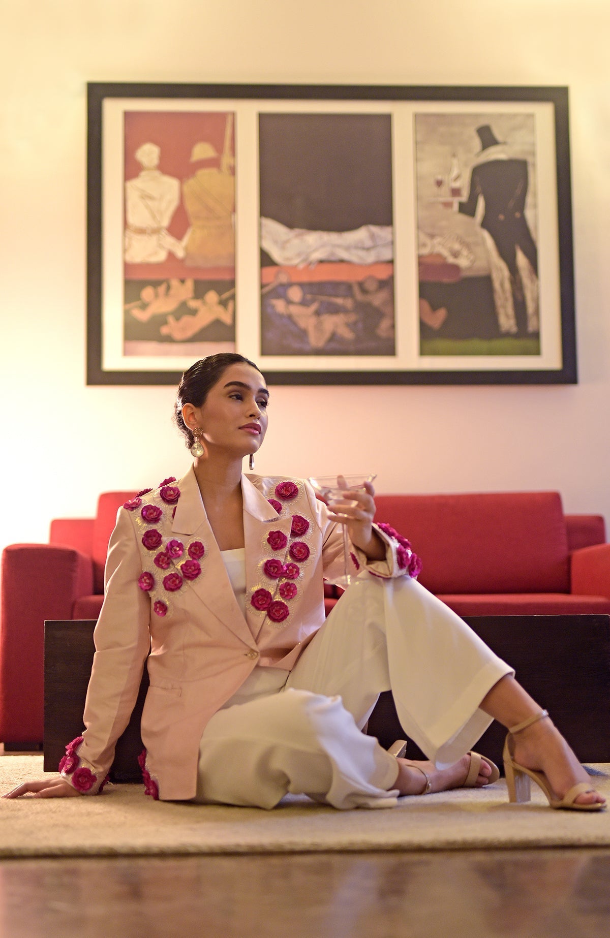 A woman in a pale pink embroidered jacket and white jumpsuit sits on the floor.
