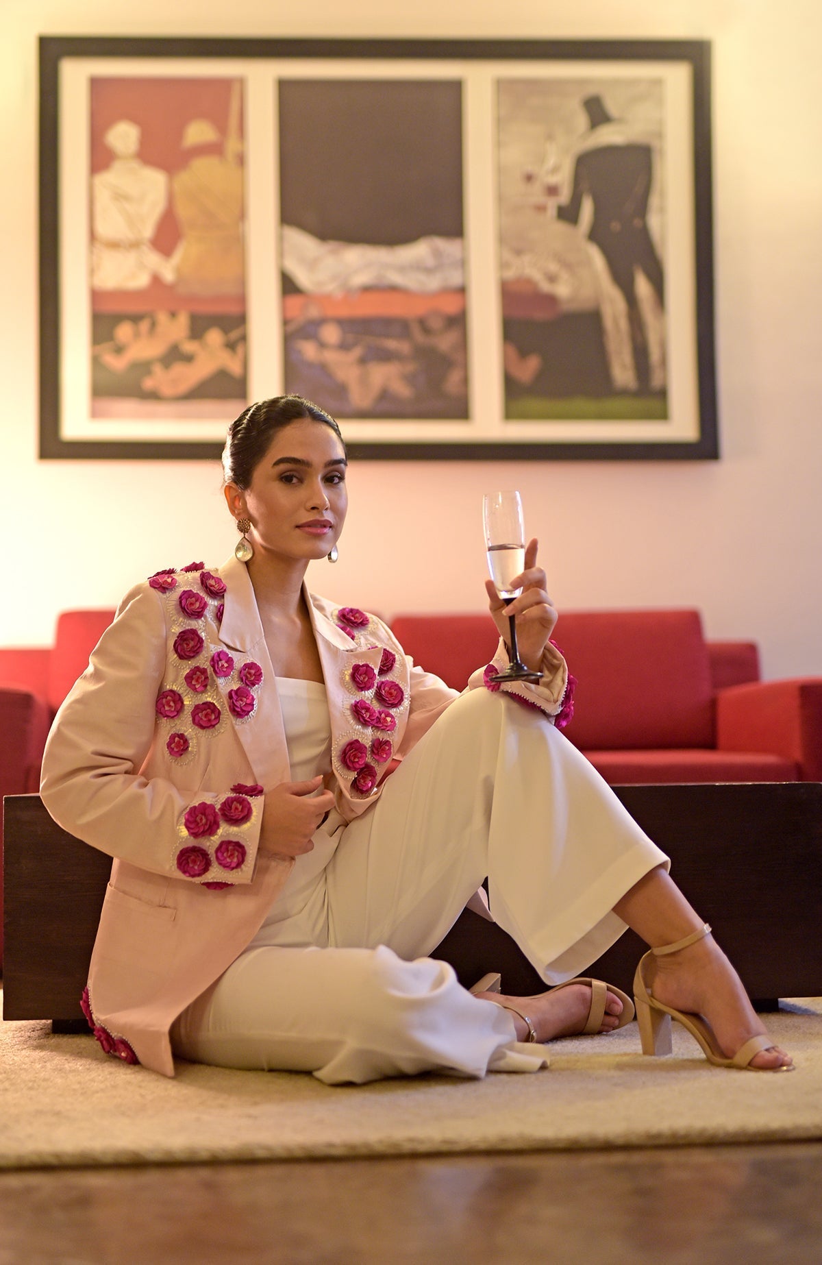A woman in a pink embroidered jacket and white jumpsuit holds a glass of champagne.