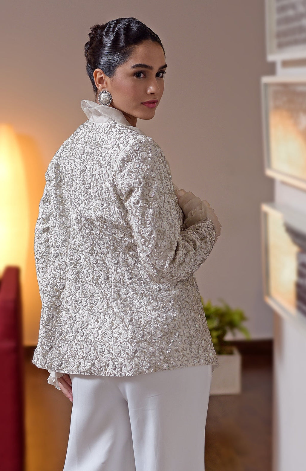 A woman in a white embroidered jacket and white jumpsuit looks over her shoulder.