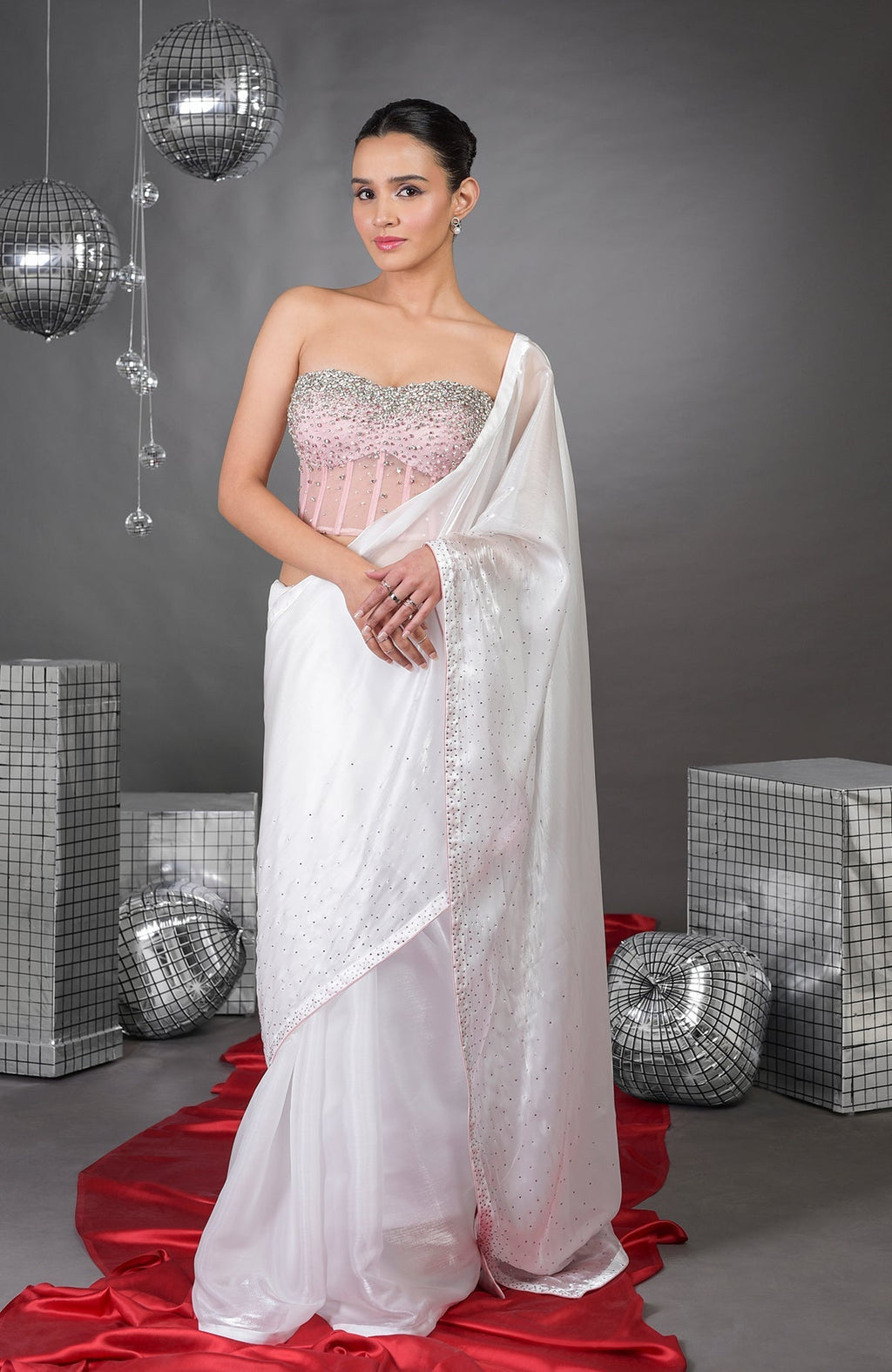 A woman models a pink corset with a white saree, standing on a red fabric.