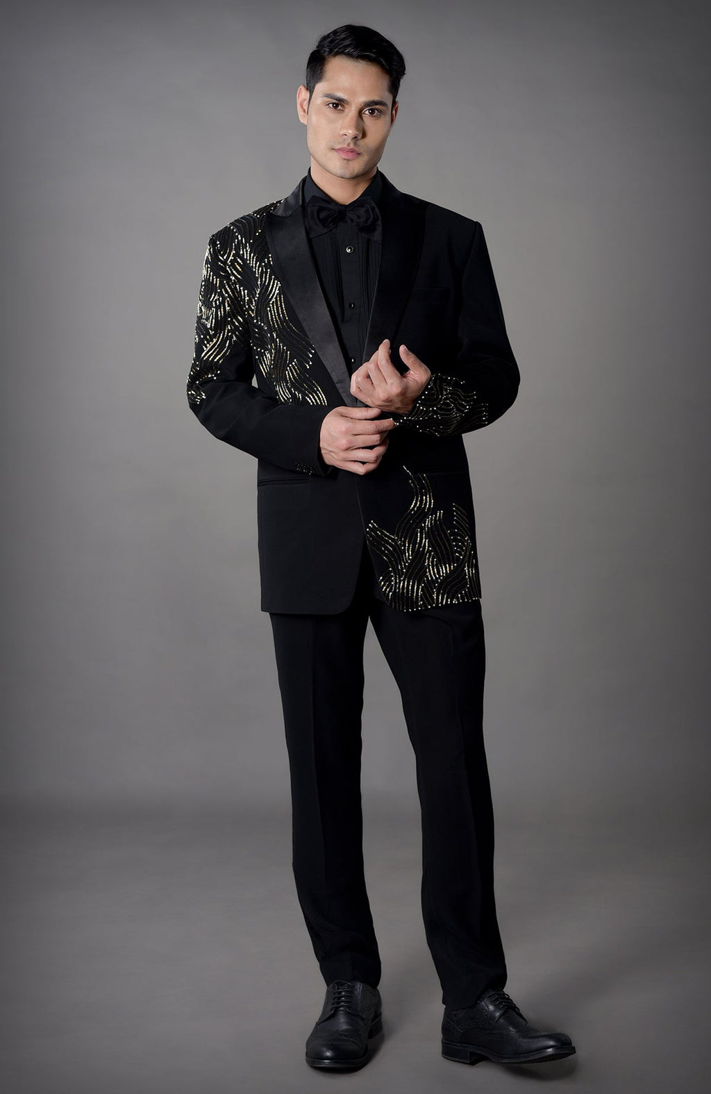 A man models a black tuxedo with gold embroidered patterns and a black bow tie.