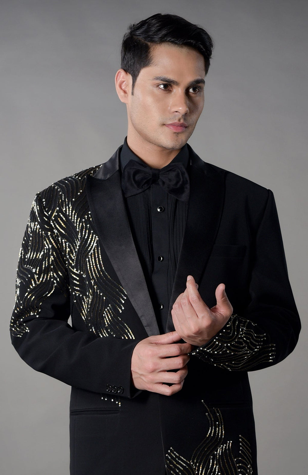 A man models a black tuxedo with gold sequin embroidery and a black bow tie.