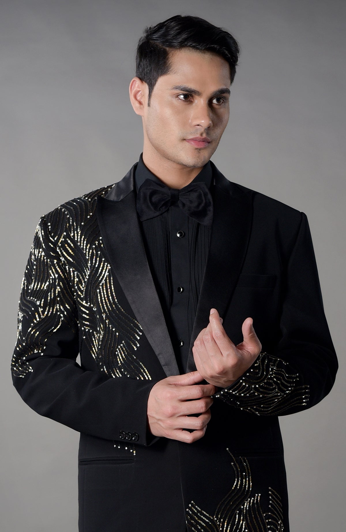 A man models a black tuxedo with gold sequin embroidery and a black bow tie.