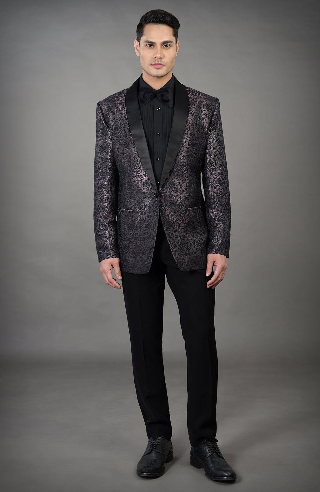 A man models a black woven brocade tuxedo with a black shirt and bow tie.
