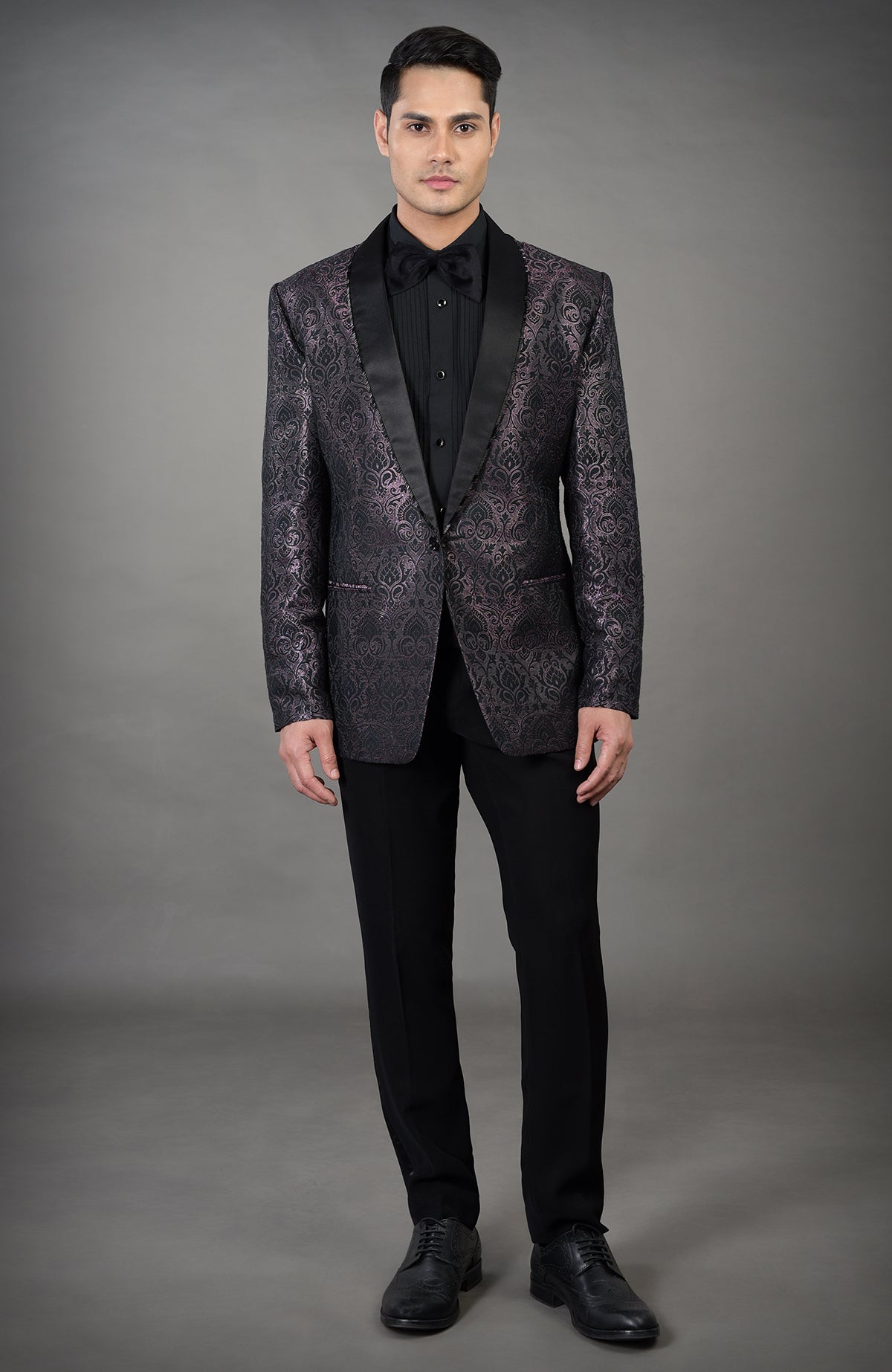 A man models a black woven brocade tuxedo with a black shirt and bow tie.